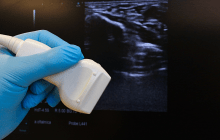 Hand holding an ultrasound probe with an ultrasound image in the background.
